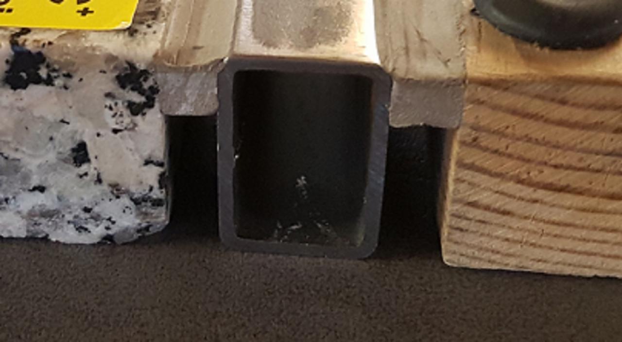 Przekrój kwadratowej rury pomiędzy fragmentem blatu z granitu z żółtą naklejką a drewnianym klockiem. Wnętrze rury jest czarne.