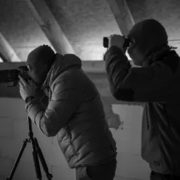 Dwóch mężczyzn w kominiarkach, jeden z aparatem na statywie, drugi z lornetką, w surowym wnętrzu budynku w Krakowie.