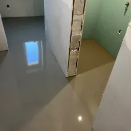 Wylany samopoziomujący podkład podłogowy w trakcie prac wykończeniowych łazienki z zielonymi płytami gipsowo-kartonowymi i oknem odbijającym światło.