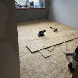 TERMON Czesław Duda - Układanie płyt OSB na podłodze w pokoju. Widoczne narzędzia: piła, młotek, odkurzacz. Surowe ściany, okno w tle. Etap prac wykończeniowych.