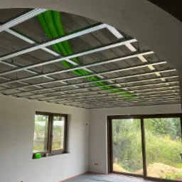 Sufit podwieszany z zielonymi rurami wentylacyjnymi w surowym wnętrzu. Widoczne okno i drzwi balkonowe z widokiem na zieleń. Podłoga zabezpieczona folią.