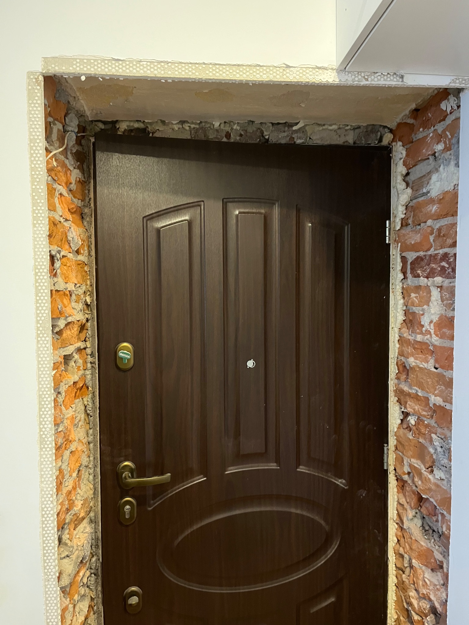 Montaż brązowych drzwi wejściowych w surowej ościeżnicy z fragmentami cegieł i pianki montażowej, widoczna listwa montażowa z otworami.