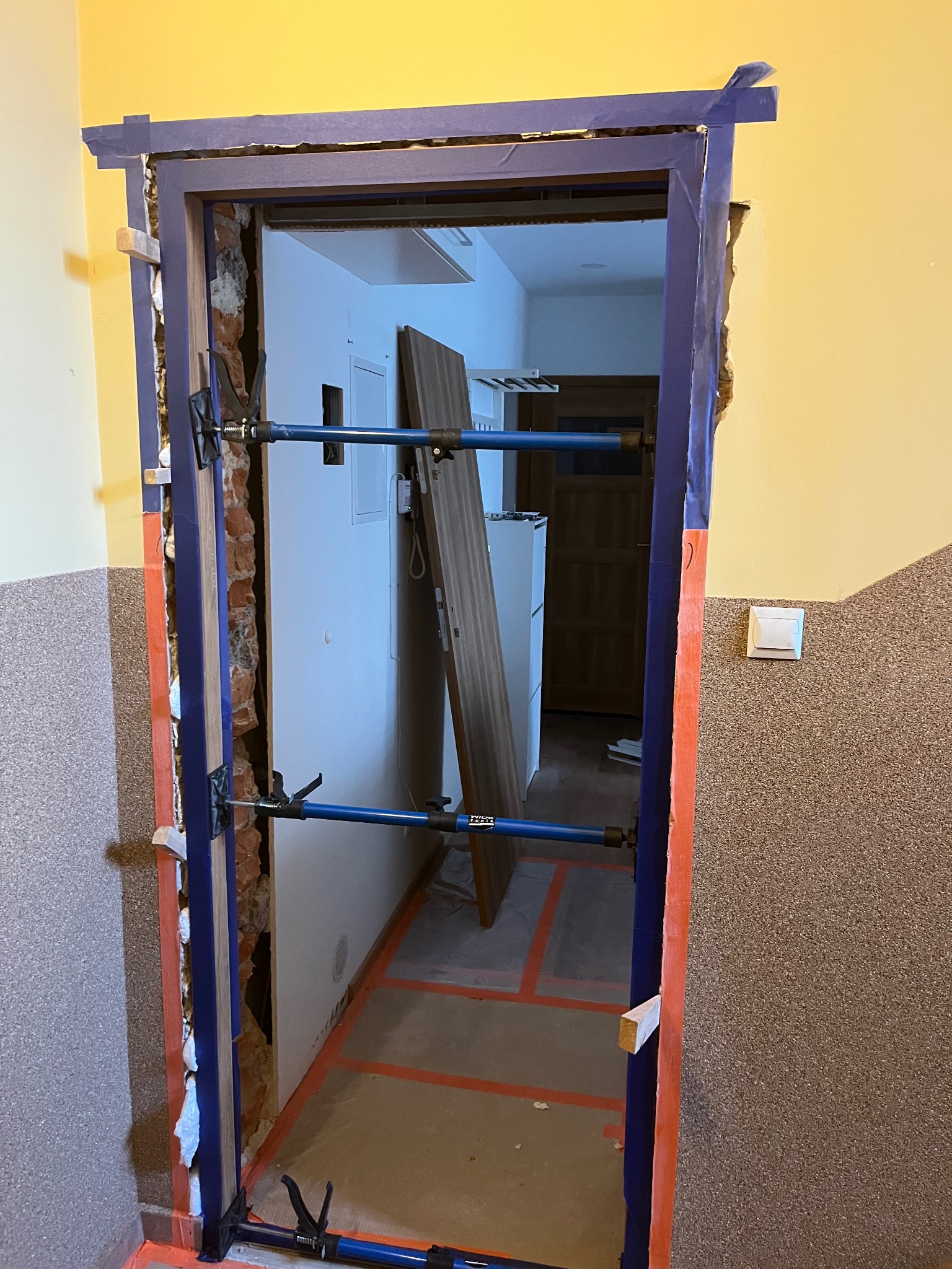 Przygotowanie do montażu ościeżnicy w łazience, zabezpieczona folią i wzmocniona rozpórkami teleskopowymi, widoczne ślady po skuciu starej futryny, w tle fragment wyposażenia łazienki.