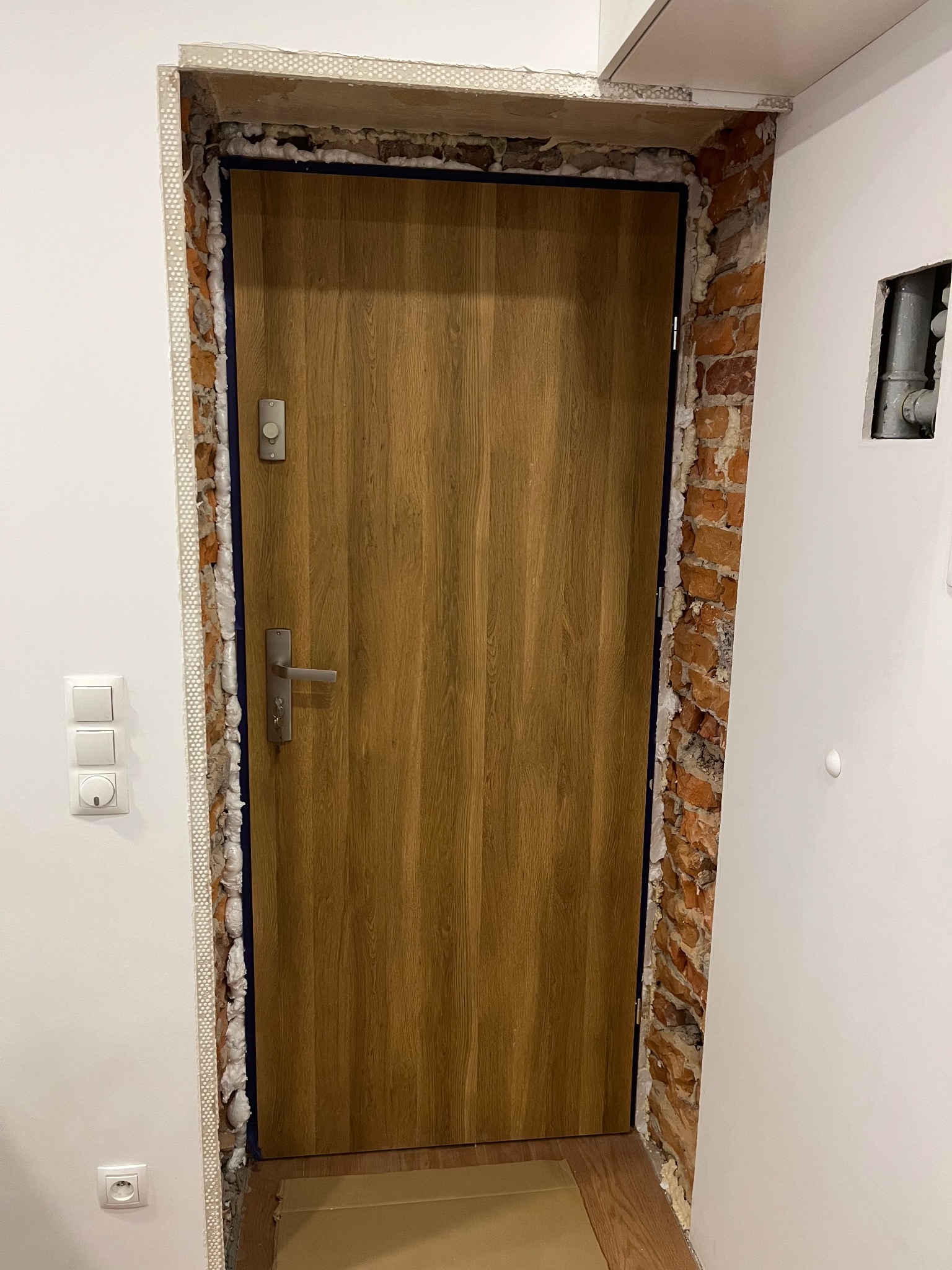 Montaż ościeżnicy drewnianej w surowym otworze z widoczną cegłą, pianką montażową i listwą perforowaną, przed wykończeniem ściany.