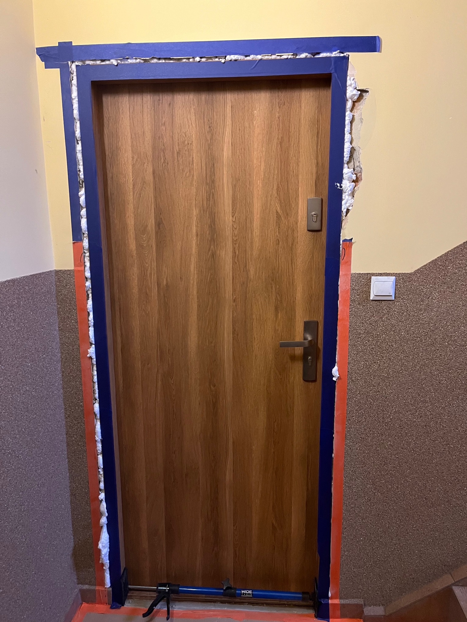 Montaż ościeżnicy drewnianej z użyciem pianki montażowej i niebieskiej taśmy malarskiej, widoczny docisk do ościeżnicy, tło stanowi ściana klatki schodowej.