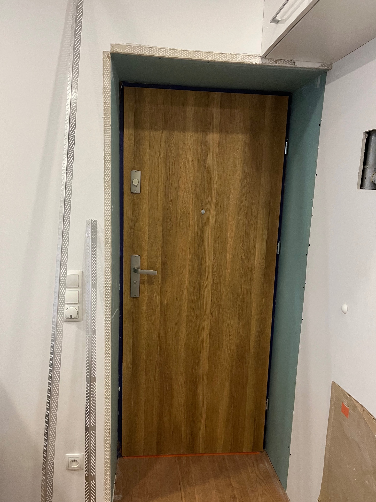 Montaż ościeżnicy drzwi wejściowych z drewnopodobną okleiną, widoczne profile wykończeniowe i płyty gipsowo-kartonowe na ścianach.