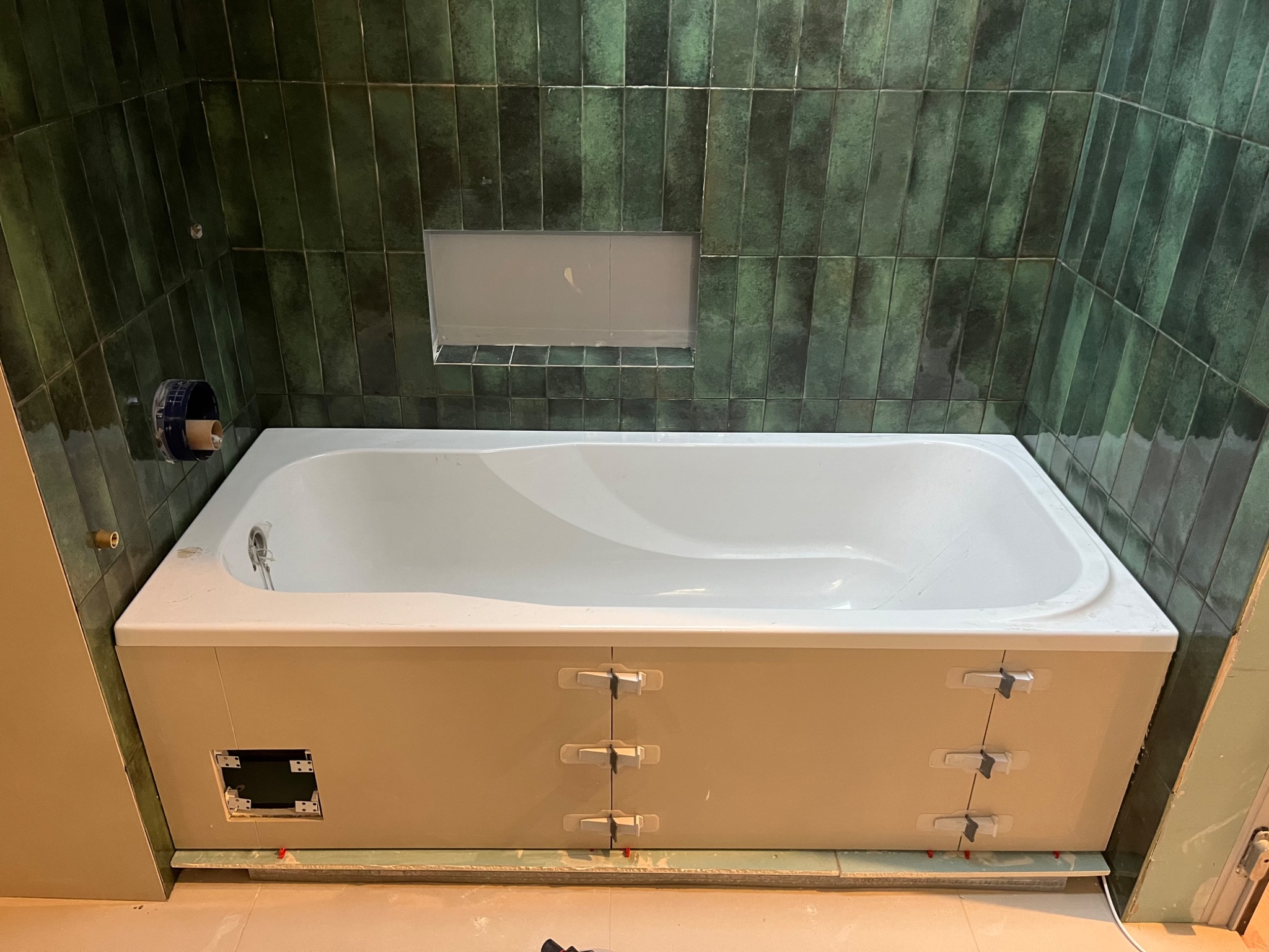 Wanna obudowana płytami, z widocznymi klipsami poziomującymi, na tle ściany wyłożonej pionowymi, zielonymi płytkami w stylu retro z wnęką.