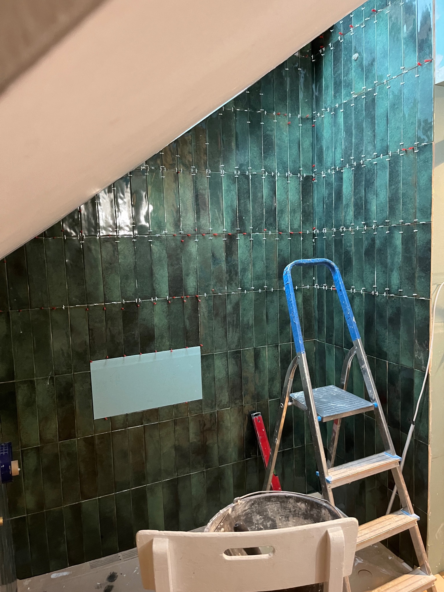 Układanie pionowych, ciemnozielonych płytek w łazience na poddaszu, widoczna drabina, poziomica i plastikowe krzyżyki dystansowe.