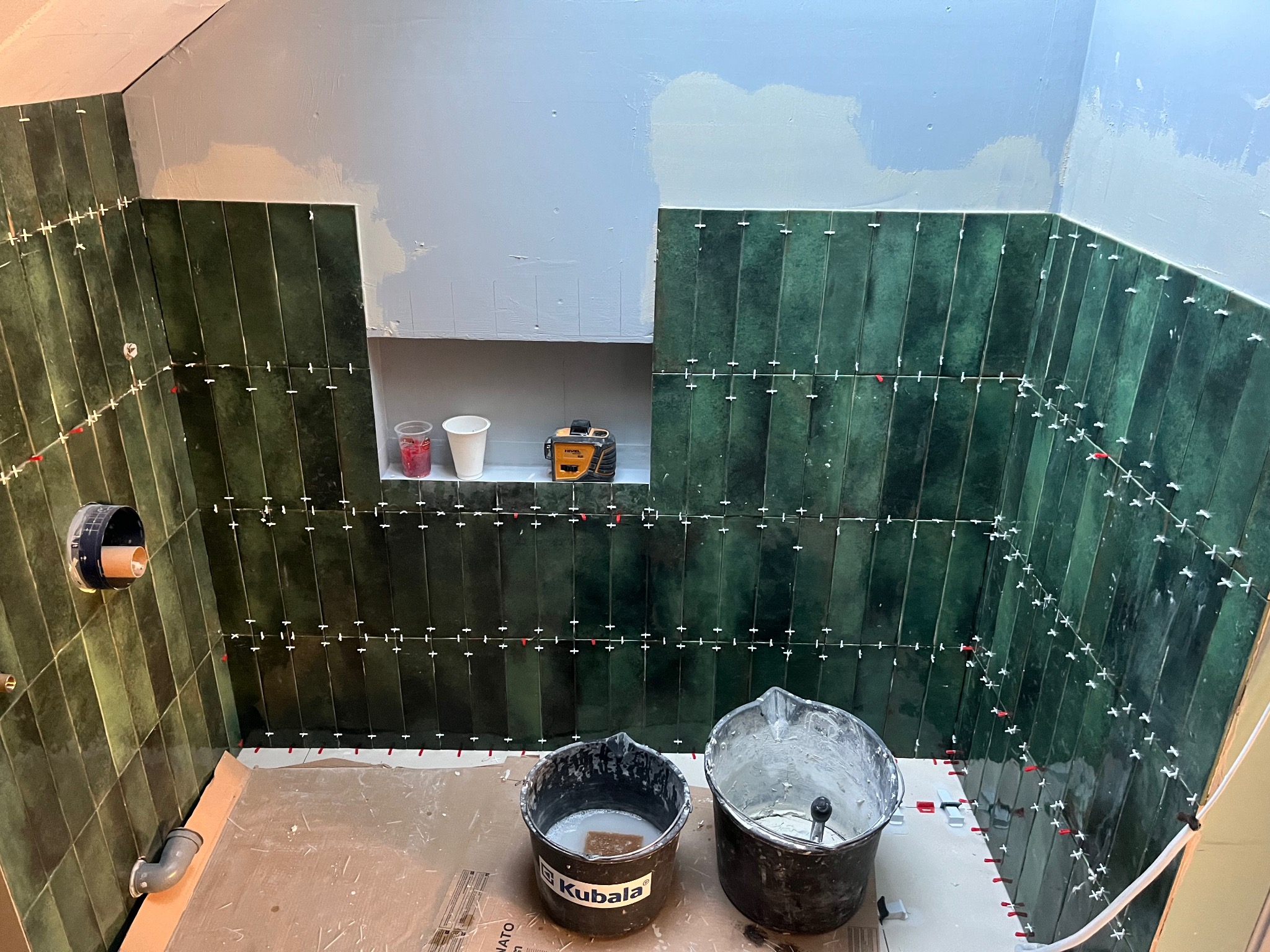 W trakcie układania pionowych, zielonych płytek w łazience; widoczne krzyżyki dystansowe, poziomica laserowa na półce, dwa wiadra z zaprawą Kubala oraz częściowo pomalowany sufit.