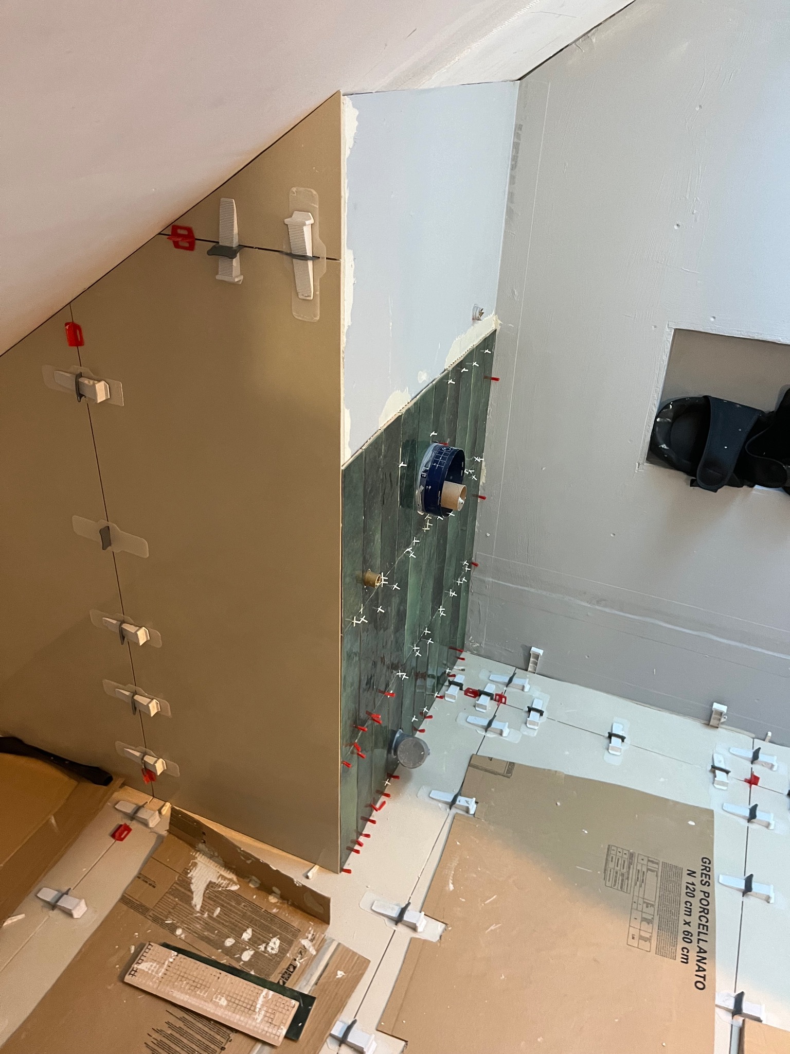 Montaż płytek gresowych w łazience na poddaszu, widoczne pionowe płytki na ścianie z systemem poziomowania, białe płytki podłogowe częściowo ułożone, kartony zabezpieczające podłogę.