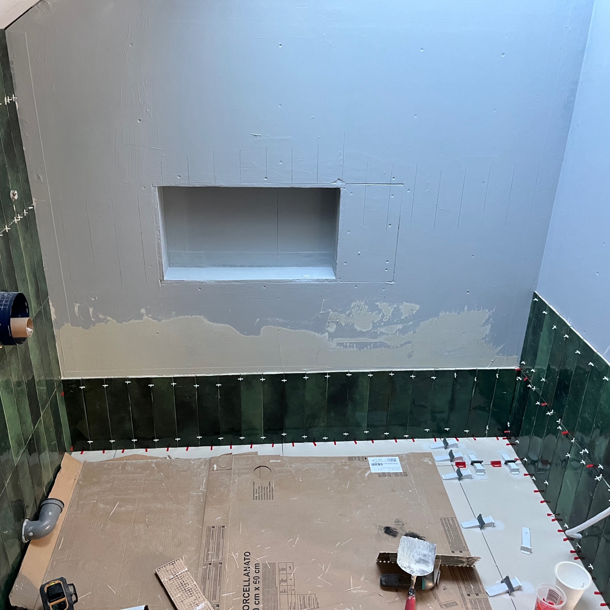 Montaż pionowych, ciemnozielonych płytek w łazience; widoczne krzyżyki dystansowe, poziomowanie laserowe, przygotowana wnęka w ścianie, częściowo położona podłoga, zabezpieczona kartonem.