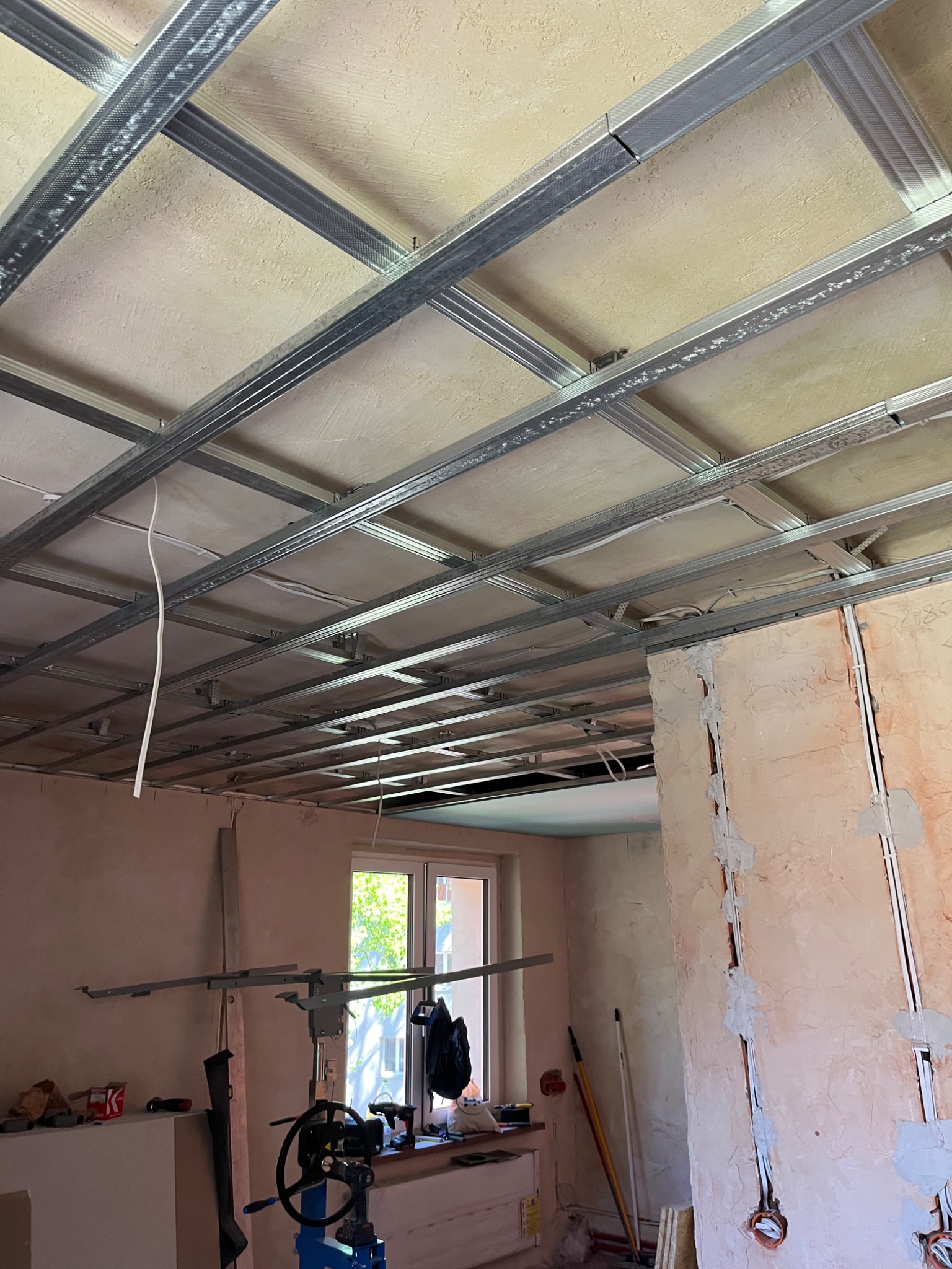 Montaż sufitu podwieszanego z płyt karton-gips. Widoczne metalowe profile i częściowo zamontowane płyty. W tle okno i ściany w trakcie prac wykończeniowych.