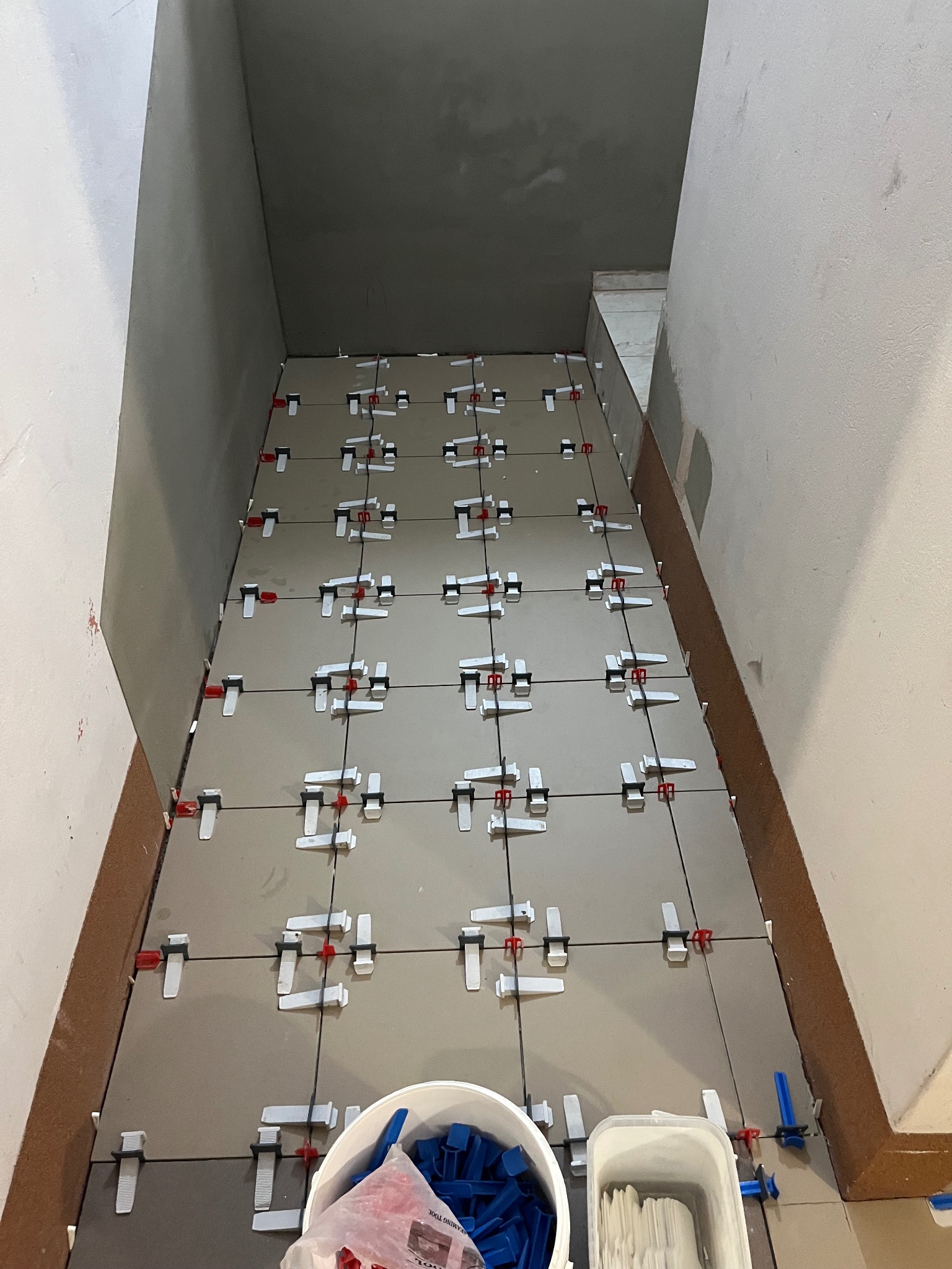 Układanie szarych płytek na podłodze w wąskim pomieszczeniu, z użyciem klipsów poziomujących. Widoczne narzędzia i wiadro z elementami montażowymi.