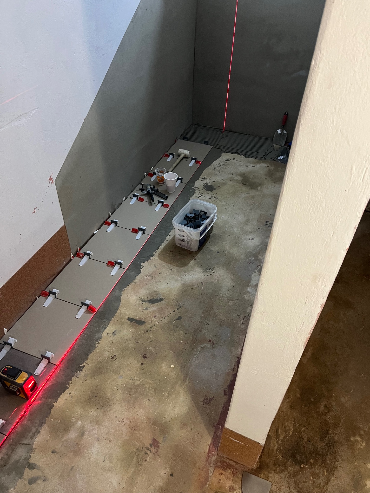 Układanie płytek w wąskim pomieszczeniu z użyciem poziomicy laserowej i systemu poziomowania. Widoczne narzędzia i materiały glazurnicze, surowy stan podłoża.