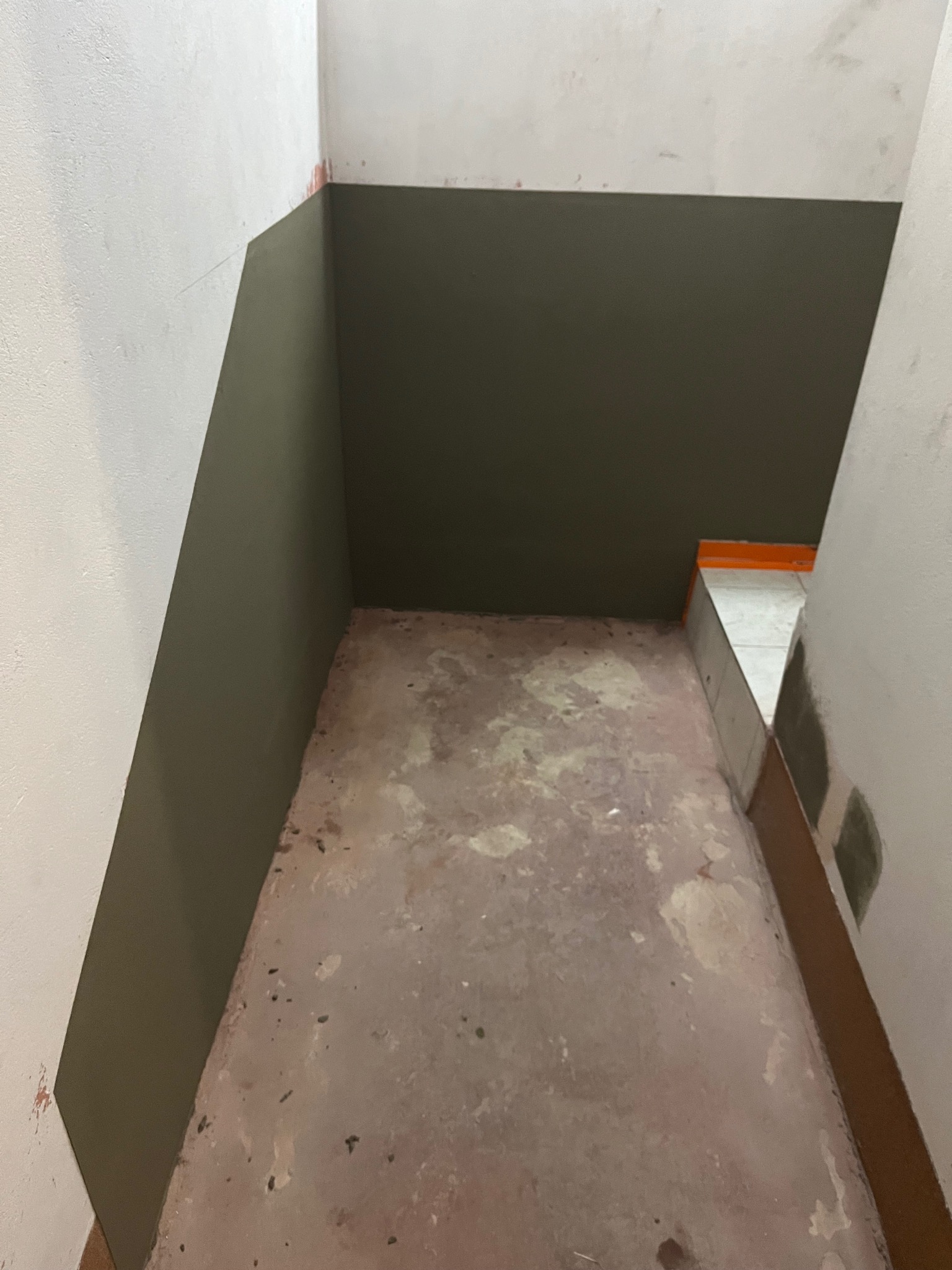 Narożnik pomieszczenia w trakcie malowania ścian na biało i oliwkowo. Widoczna surowa podłoga i fragment wykończonej podłogi z płytkami. Prace wykończeniowe.