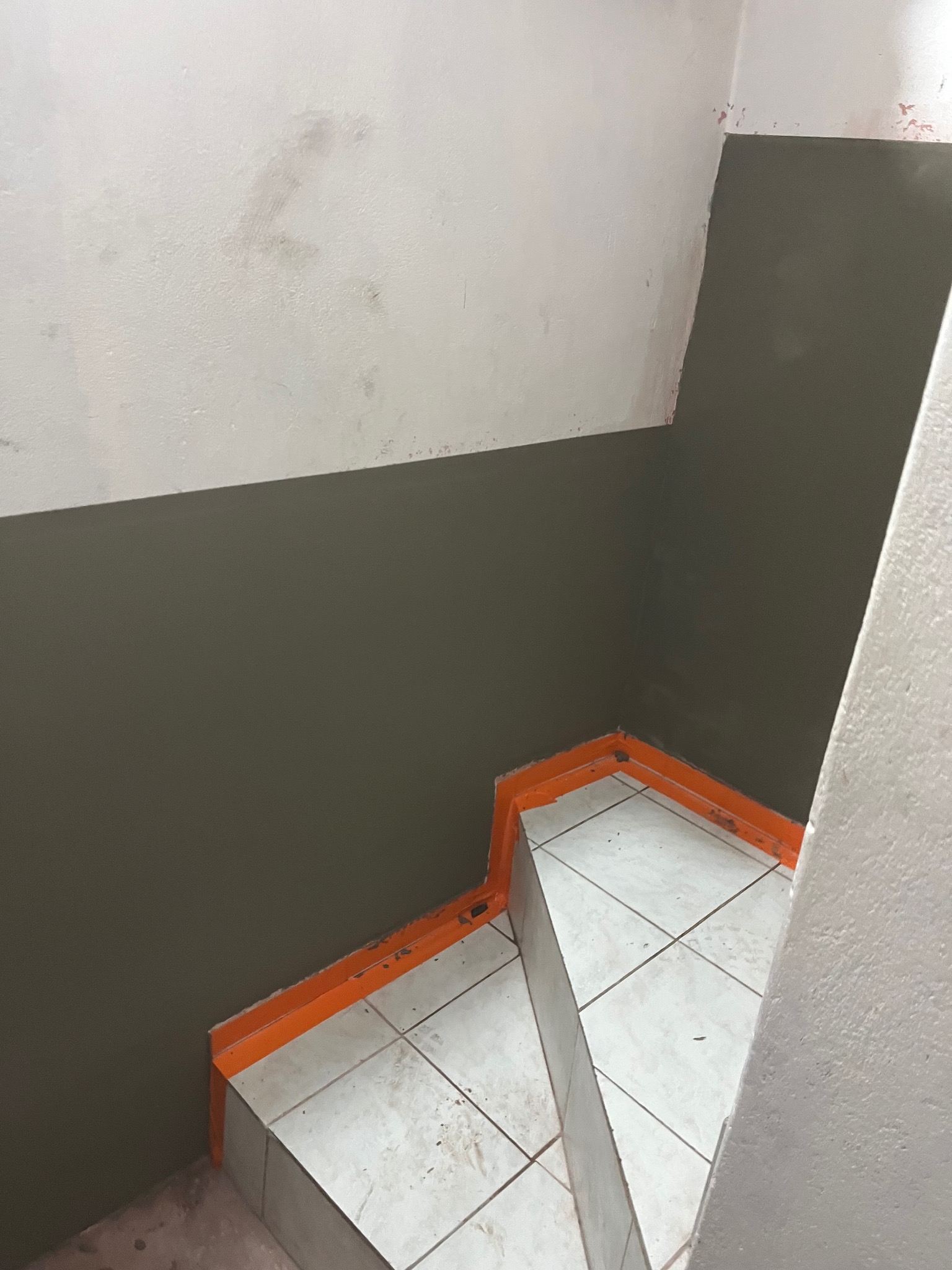 Świeżo pomalowana ściana w dwóch odcieniach szarości nad schodami wyłożonymi białymi płytkami, zabezpieczonymi pomarańczową taśmą malarską.