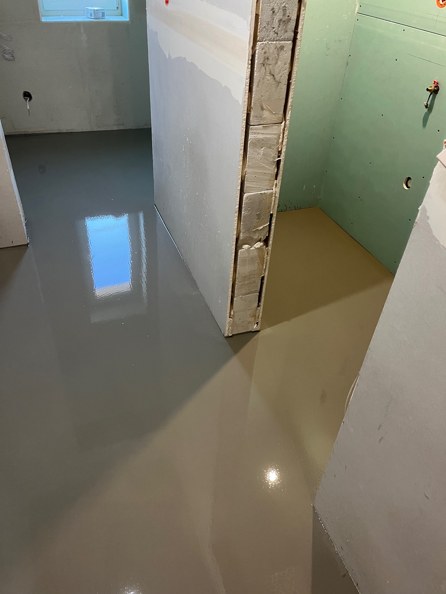 Wylany samopoziomujący podkład podłogowy w trakcie prac wykończeniowych łazienki z zielonymi płytami gipsowo-kartonowymi i oknem odbijającym światło.