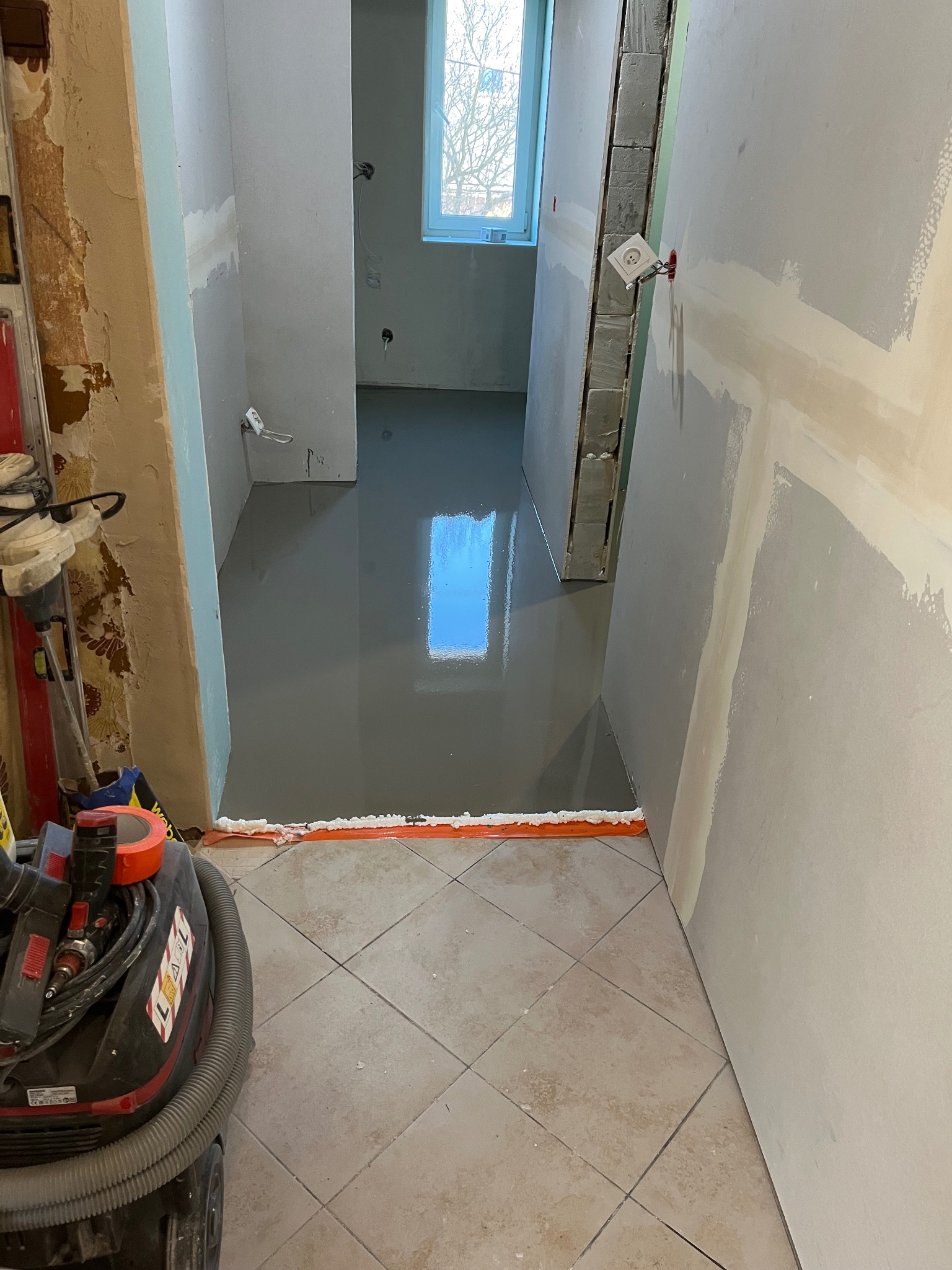 Wylewka samopoziomująca w łazience w trakcie remontu, widoczna granica wylewki z pomarańczową taśmą, narzędzia i płytki podłogowe. Ściany przygotowane do dalszych prac.