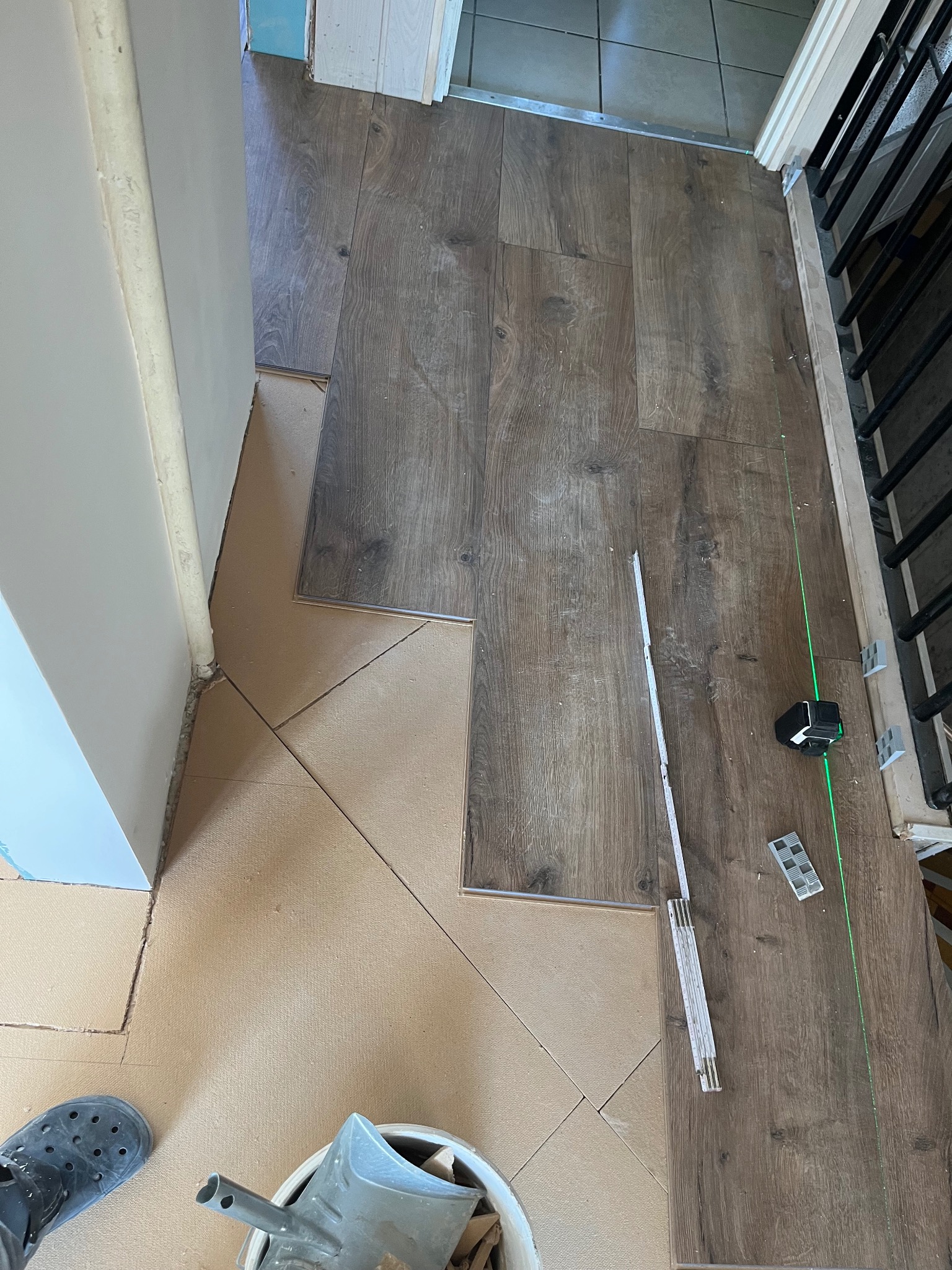 Układanie paneli podłogowych w trakcie remontu. Widoczny laser krzyżowy, miarka składana i fragment podłoża z brązowych płytek. Częściowo ułożone panele w odcieniach brązu.