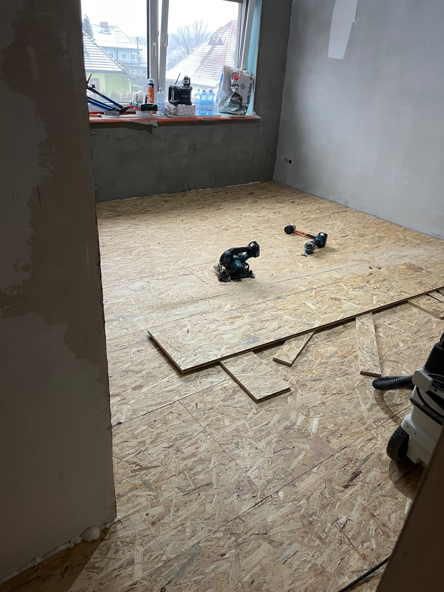 Układanie płyt OSB na podłodze w pokoju. Widoczne narzędzia: piła, młotek, odkurzacz. Surowe ściany, okno w tle. Etap prac wykończeniowych.