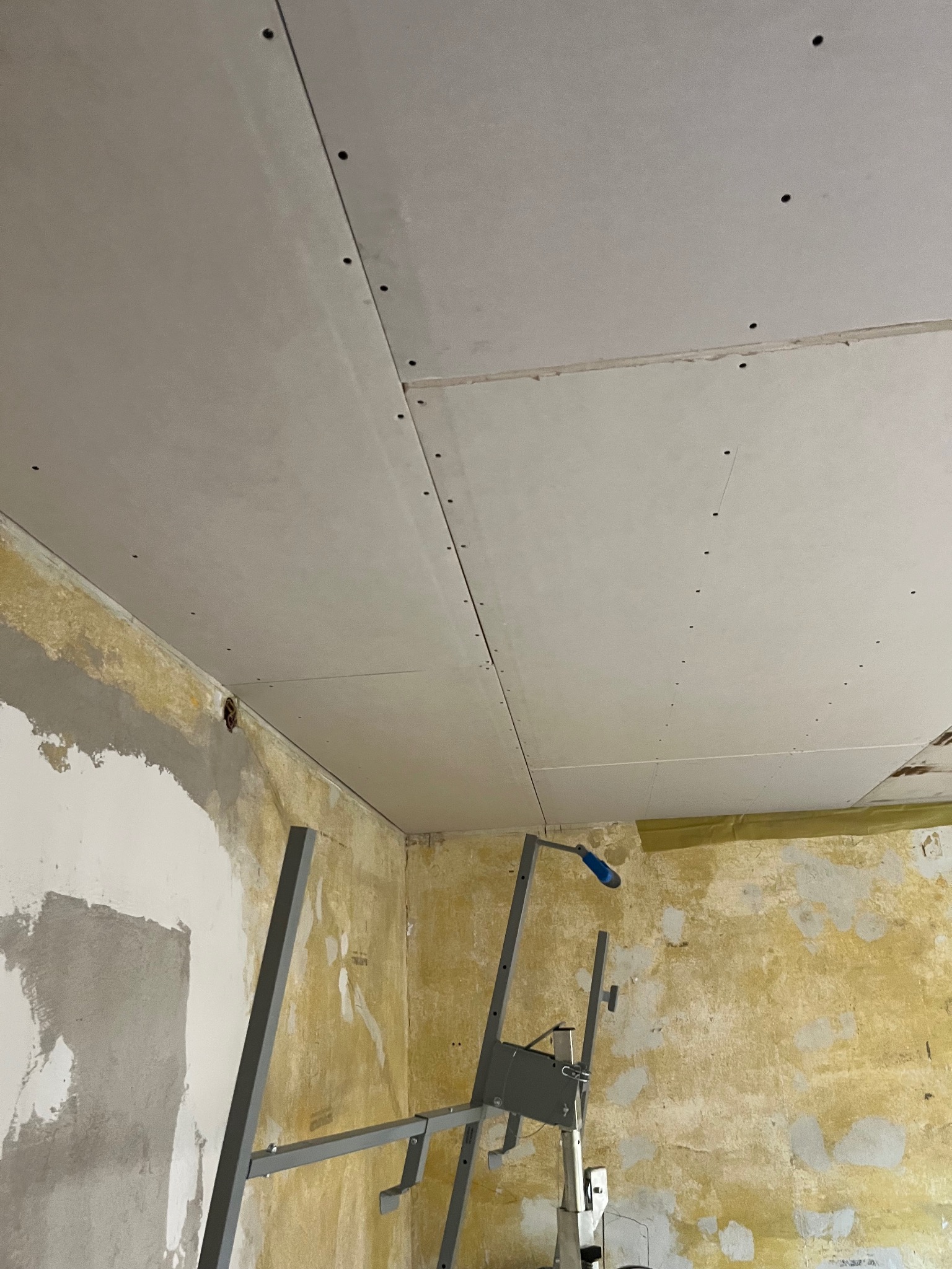 Montaż sufitu z płyt gipsowo-kartonowych w pomieszczeniu z widoczną starą powłoką ścian, wsparty na metalowym stelażu. Widoczne ślady po wkrętach.