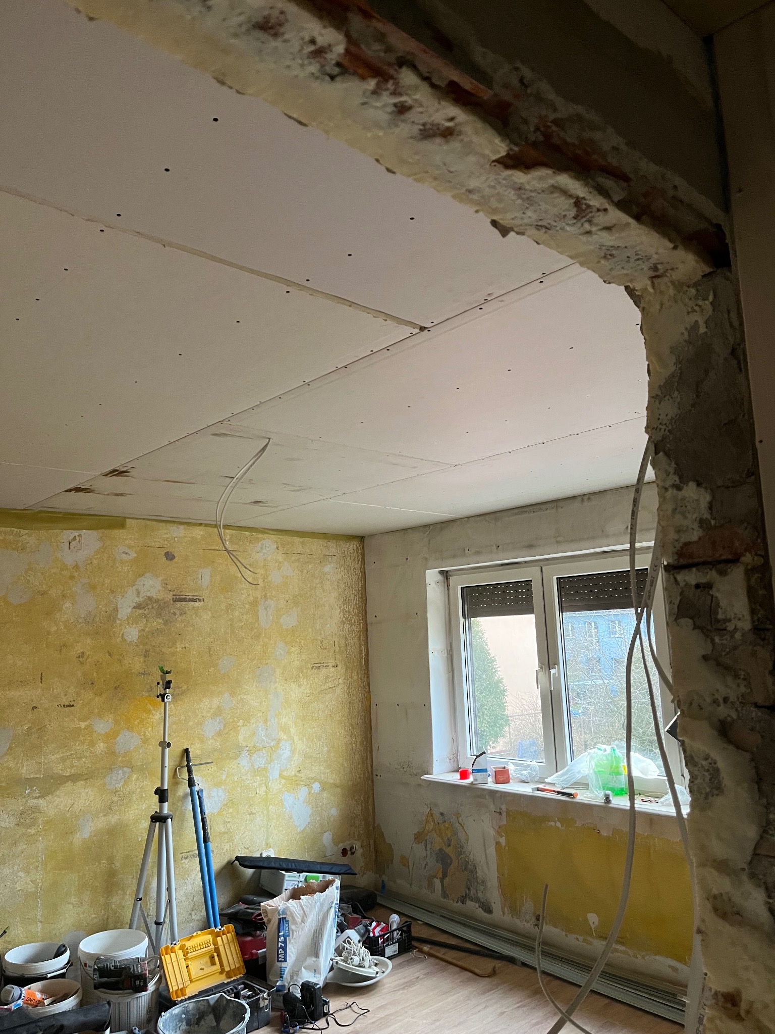 Remont pokoju: sufit z płyt karton-gips, ściany z resztkami farby, narzędzia, okno z roletami. Widoczne kable i elementy konstrukcyjne.