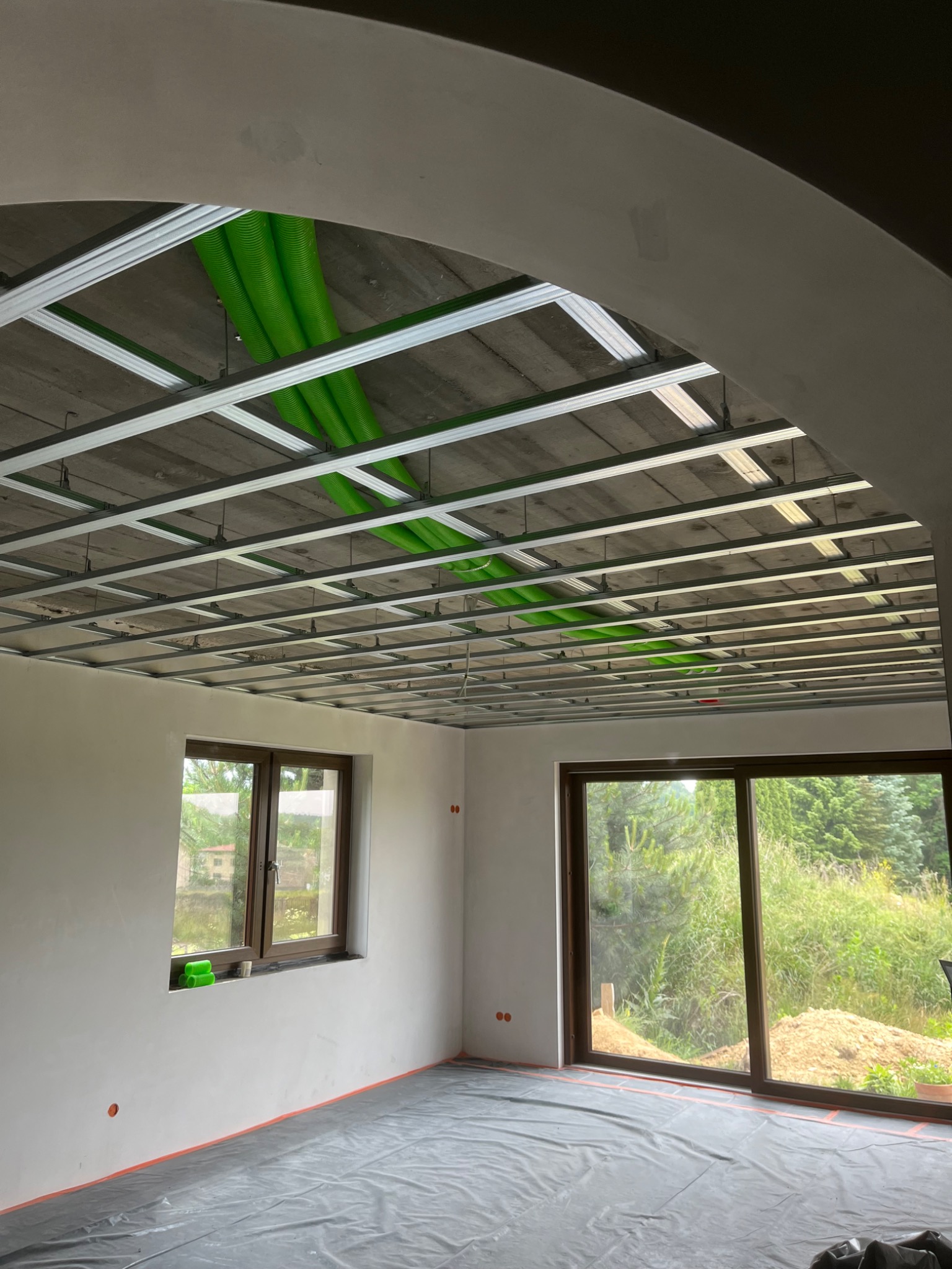 Sufit podwieszany z zielonymi rurami wentylacyjnymi w surowym wnętrzu. Widoczne okno i drzwi balkonowe z widokiem na zieleń. Podłoga zabezpieczona folią.