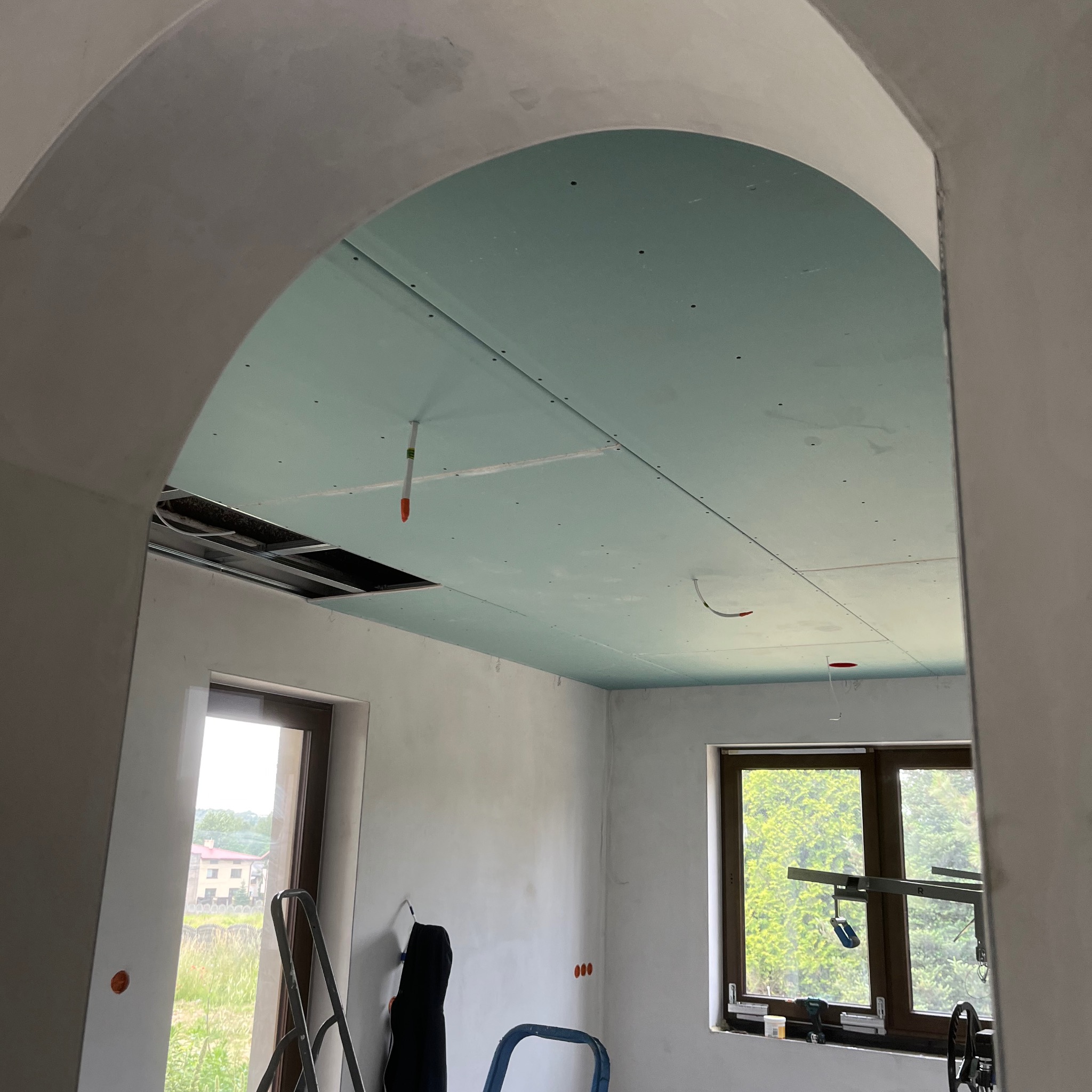 Montaż sufitu podwieszanego z płyty GK w pomieszczeniu z oknami. Widoczne profile, płyty w kolorze zielonym oraz elementy instalacji elektrycznej.