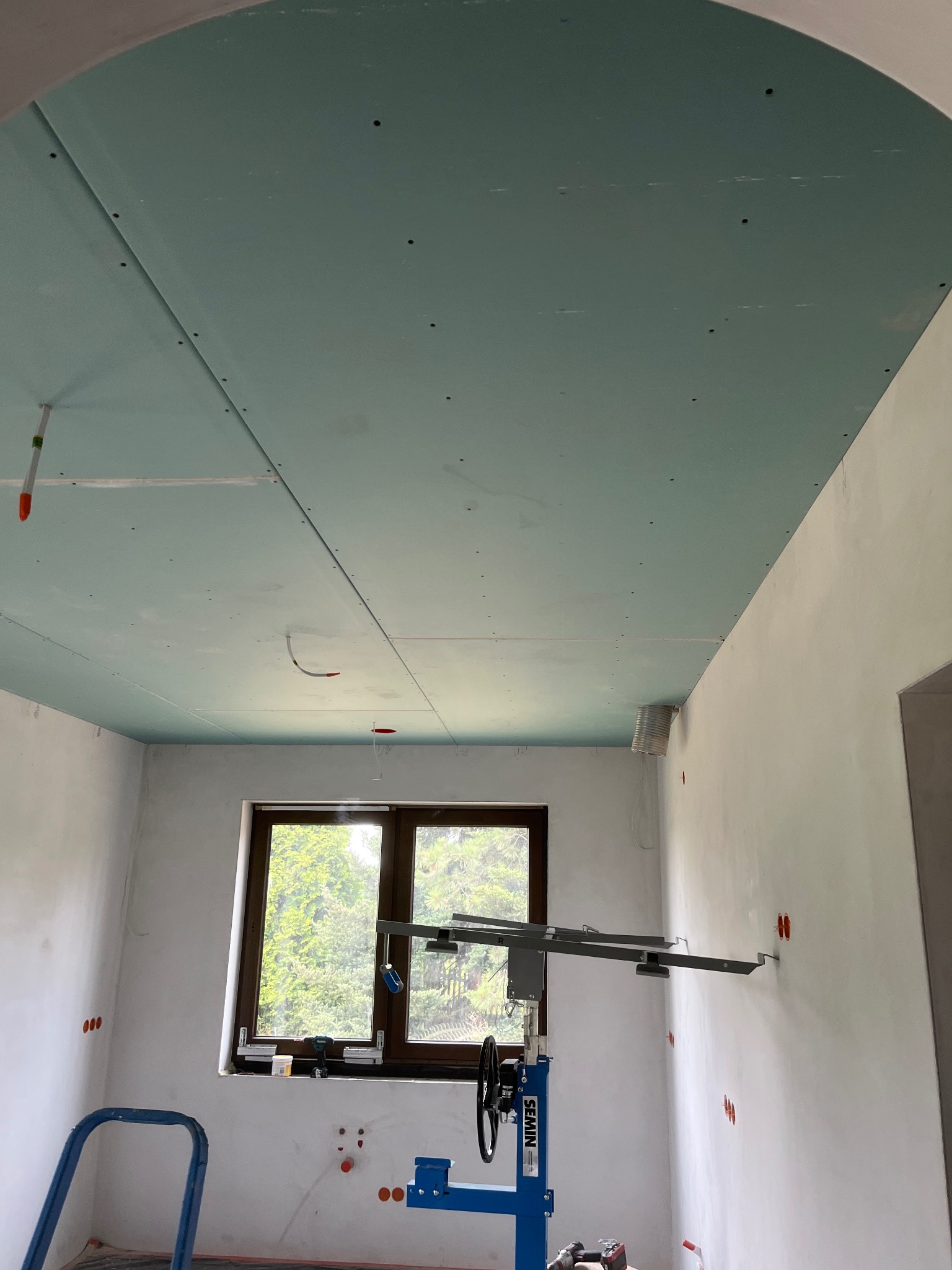 Montaż sufitu z płyty karton-gips w pomieszczeniu z oknem. Widoczny podnośnik do płyt i wystające kable. Sufit w kolorze delikatnej zieleni.