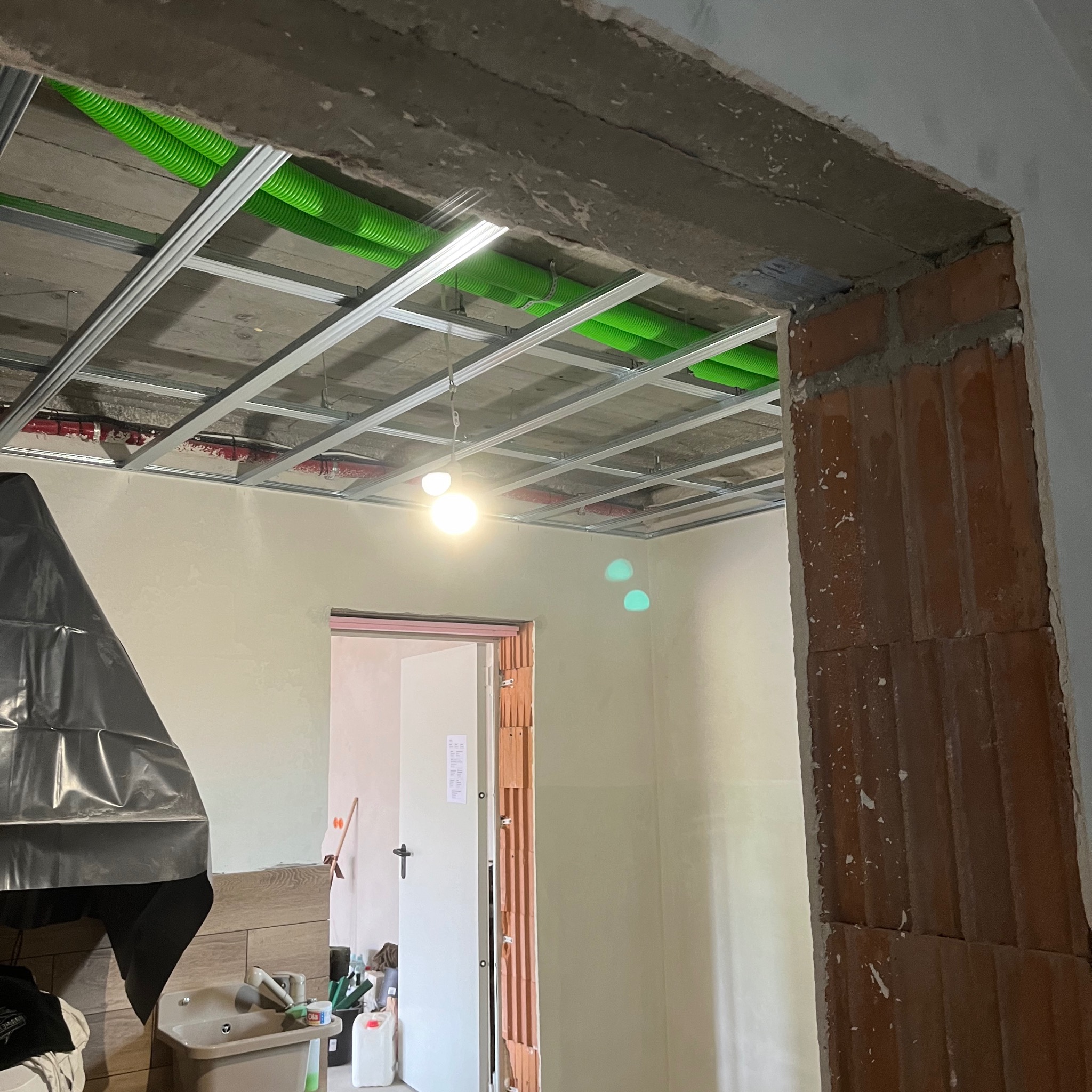 Sufit podwieszany z metalową konstrukcją i zielonymi rurami instalacyjnymi, widok na wnętrze w trakcie remontu, fragment ściany z cegły.