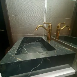 Nowoczesna umywalka z czarnego marmuru z złotą armaturą, odbijająca się w lustrze na tle teksturowanej ściany.