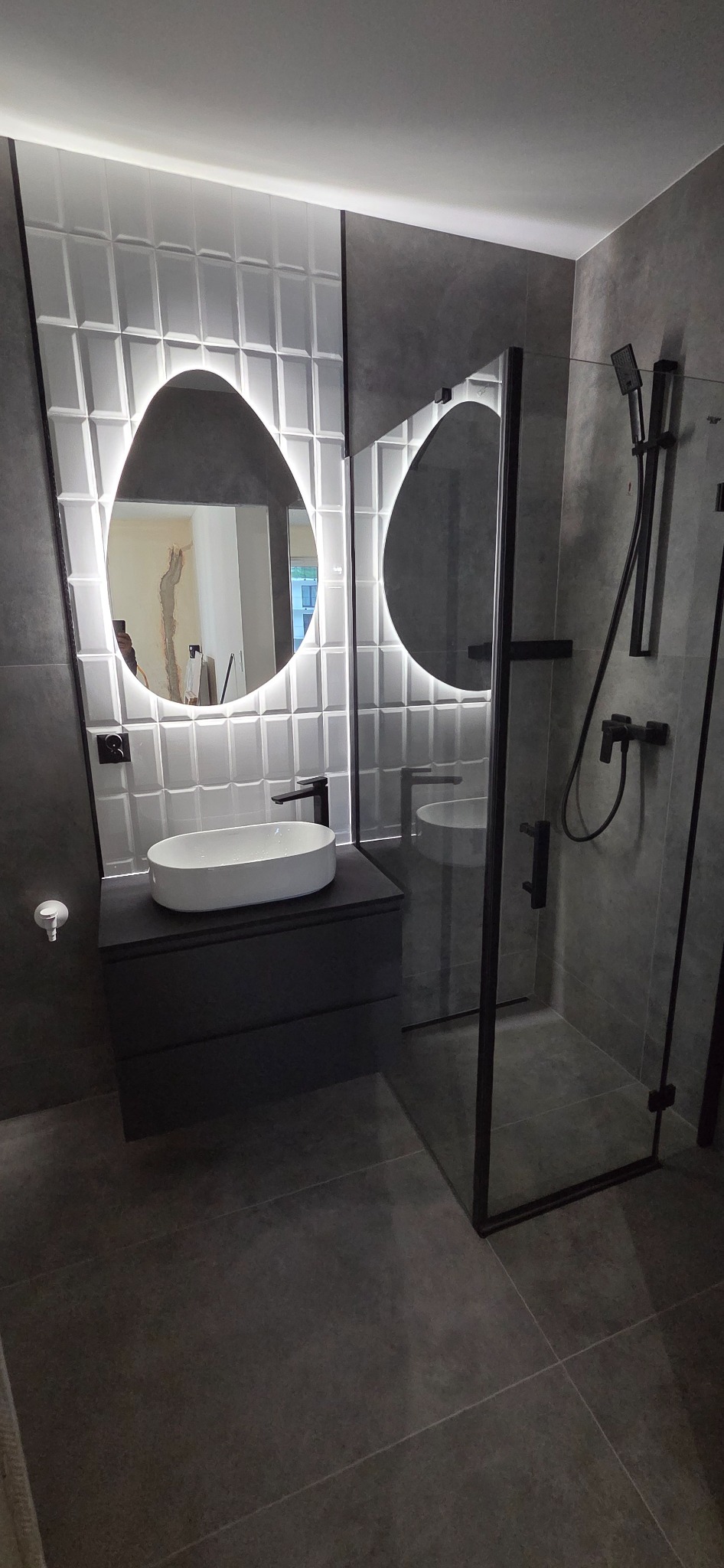 Nowoczesna łazienka w szarościach z podświetlanym lustrem o nieregularnym kształcie, kabiną prysznicową typu walk-in i umywalką nablatową. Widoczne płytki 3D na ścianie.