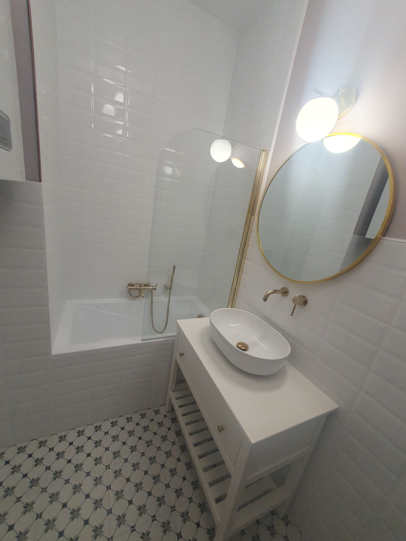 Nowoczesna łazienka po remoncie: białe płytki z fakturą, owalna umywalka na szafce, okrągłe lustro w złotej ramie i dekoracyjna podłoga w geometryczny wzór.