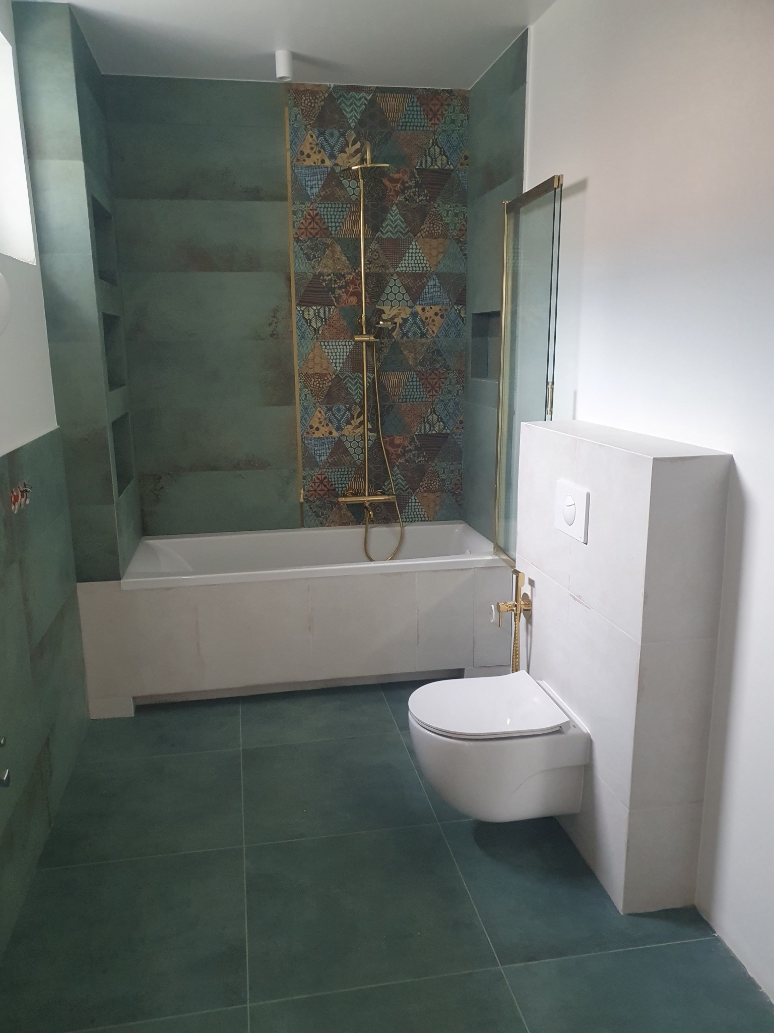 Nowoczesna łazienka z wanną, kabiną prysznicową i podwieszaną toaletą. Uwagę przykuwają zielone płytki i złota armatura, tworzące elegancki kontrast.