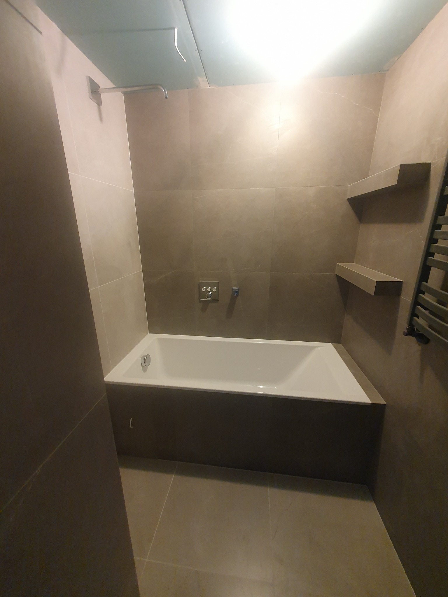 Nowoczesna łazienka z wanną w zabudowie, wykończona płytkami w odcieniach szarości. Widoczne półki ścienne i grzejnik drabinkowy. Minimalistyczny design.