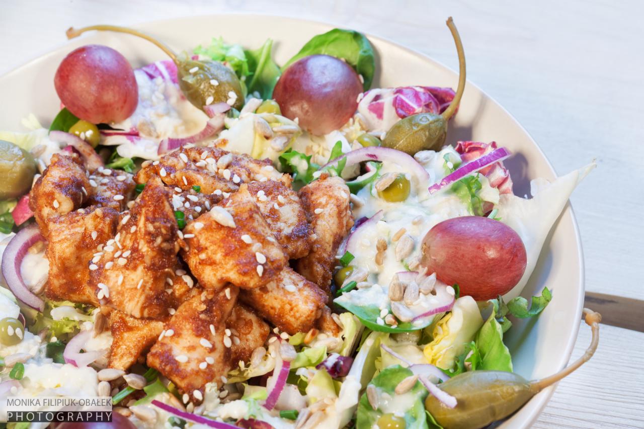 Apetyczna sałatka z grillowanym kurczakiem, czerwonymi winogronami, kaparami i czerwoną cebulą, posypana sezamem, podana w białej misce.