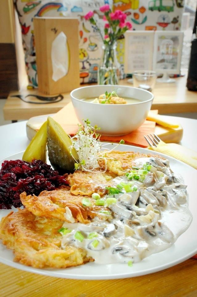 Zestaw obiadowy: placki ziemniaczane z sosem grzybowym, buraczki, ogórek kiszony i zupa krem w białej misce, całość udekorowana kiełkami i posiekanym szczypiorkiem, podana na jasnym drewnianym stole.