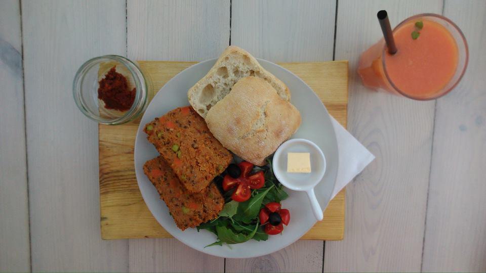 Kompozycja wegetariańskiego lunchu: pasztet warzywny, pieczywo, sałatka z pomidorami i oliwkami, masło i sok na drewnianej desce na tle białych desek.