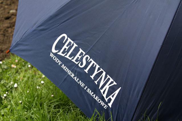 Niebieski parasol z białym nadrukiem 'Celestynka wody mineralne i smakowe' na tle trawy i ziemi.