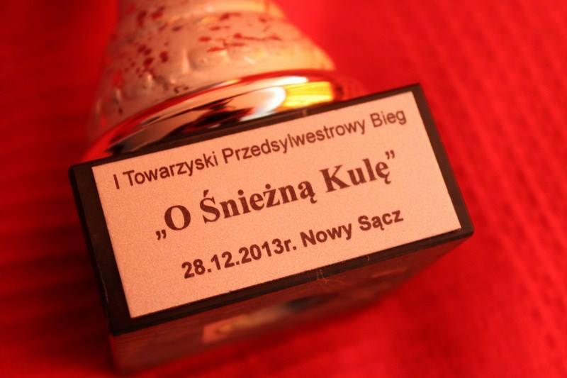 Statuetka z napisem 'I Towarzyski Przedsylwestrowy Bieg 'O Śnieżną Kulę' 28.12.2013r. Nowy Sącz' na czerwonym tle.