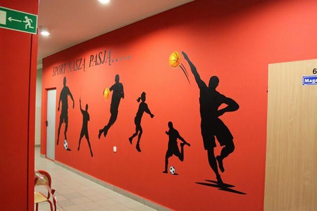 Korytarz z czerwoną ścianą ozdobioną czarnymi sylwetkami sportowców uprawiających koszykówkę i piłkę nożną oraz napisem 'Sport Naszą Pasją'.