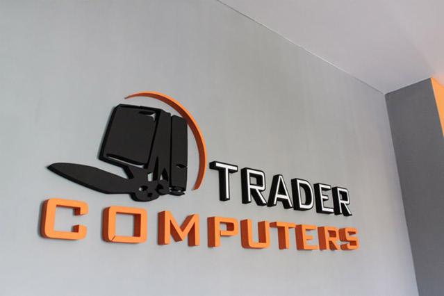 Eleganckie logo firmy komputerowej 'Trader Computers' wykonane z czarnego i pomarańczowego tworzywa, umieszczone na szarej ścianie w biurze.