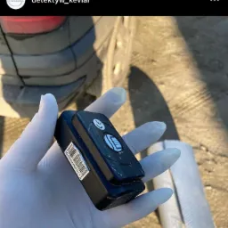 Dłoń w białej rękawiczce trzyma czarny lokalizator GPS z kodem kreskowym, w tle fragment opony samochodu i jasny zw&oacute;j materiału.