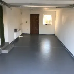 Gładka, szara posadzka w garażu z białymi ścianami i brązowymi drzwiami wejściowymi, widoczny grzejnik i instalacja wodna, okno z widokiem na zieleń na zewnątrz.