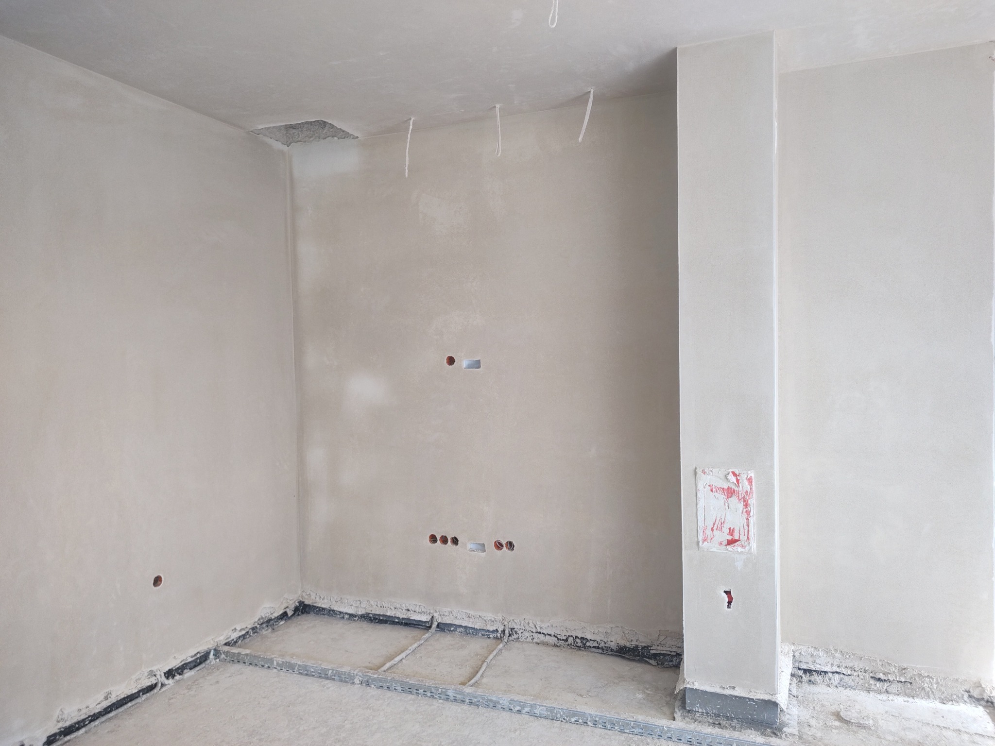 Wnętrze pokoju w trakcie prac wykończeniowych: ściany pokryte świeżym tynkiem, widoczne przewody elektryczne i listwy przypodłogowe. Otwór w suficie.