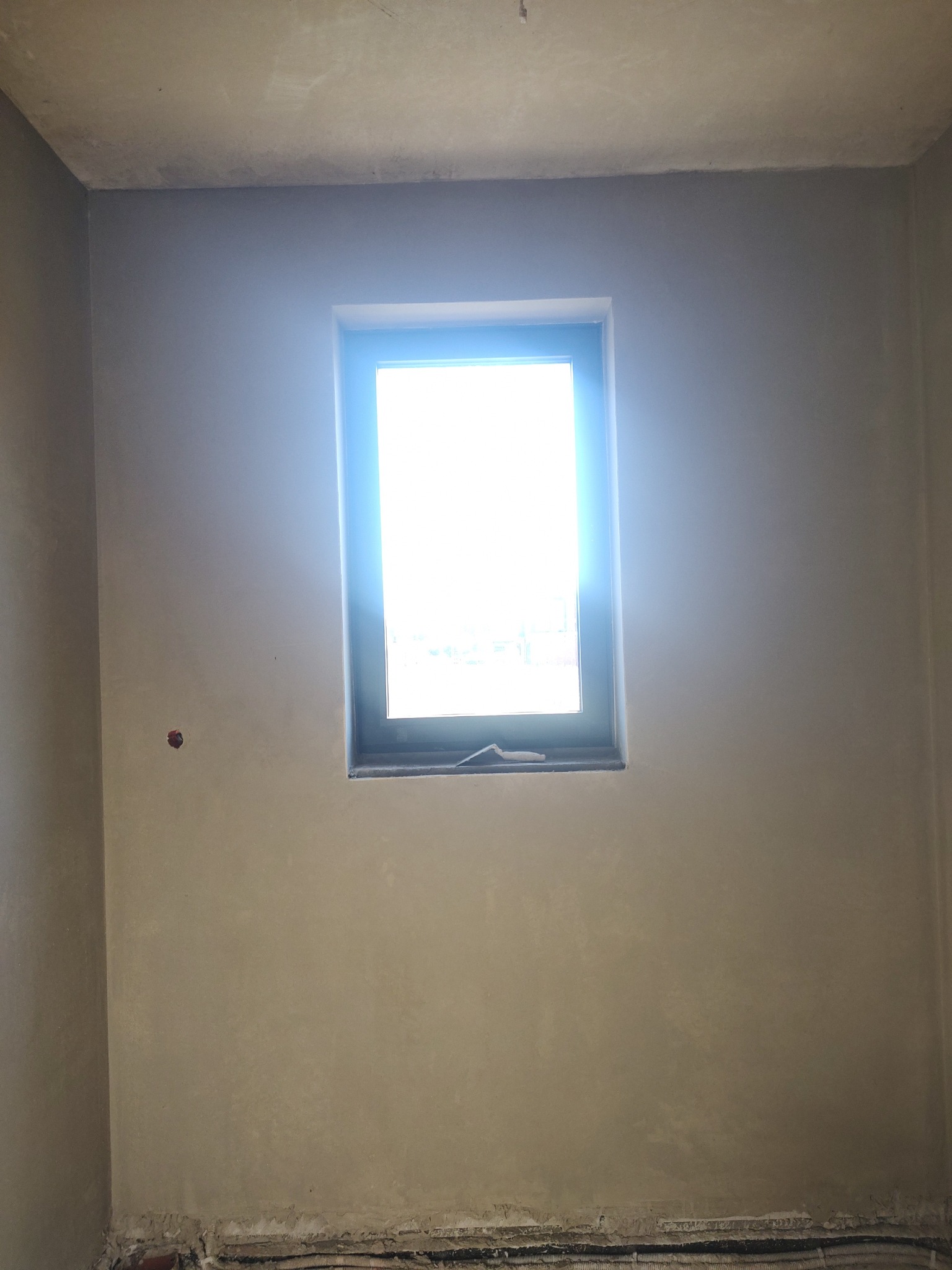 Pokój po nałożeniu tynku z oknem, widoczny fragment podłogi. Ściany i sufit w jasnym odcieniu beżu, świeżo po nałożeniu tynku.