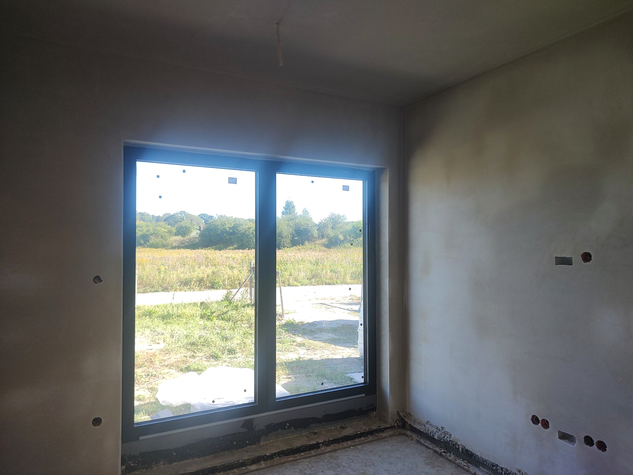 Pokój po tynkowaniu maszynowym: ściany pokryte świeżym tynkiem, widoczne otwory na gniazdka elektryczne i duże okno z widokiem na zieleń na zewnątrz.