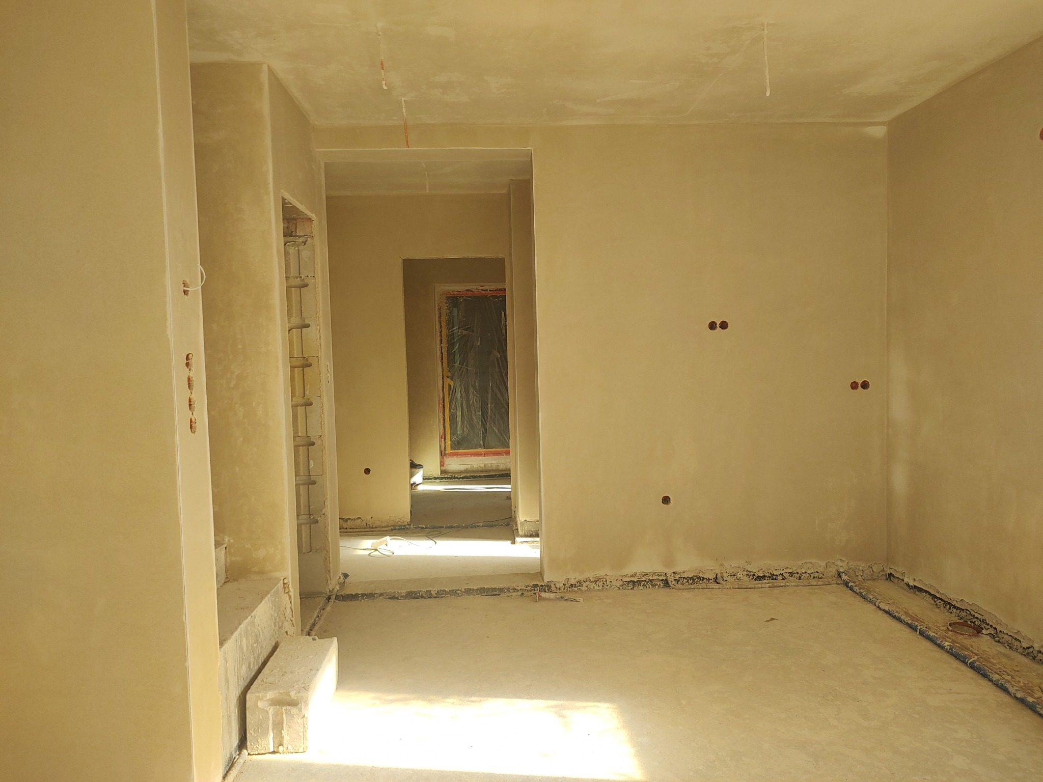 Wnętrze po nałożeniu gładzi, widoczne otwory na gniazdka elektryczne i rury w podłodze. Ściany i sufit w jasnym odcieniu beżu. W tle widoczne kolejne pomieszczenia.
