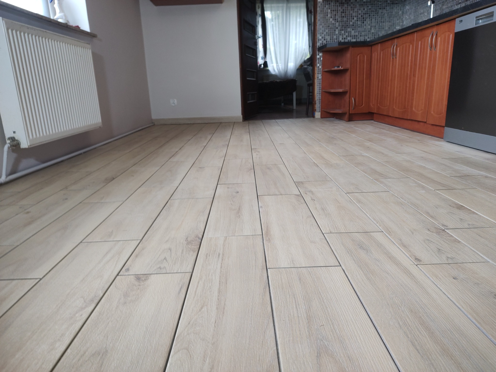 Jasne, drewnopodobne płytki podłogowe ułożone wzdłużnie, widoczne w salonie z fragmentem białego grzejnika i kuchni w tle. Ujęcie z niskiej perspektywy.