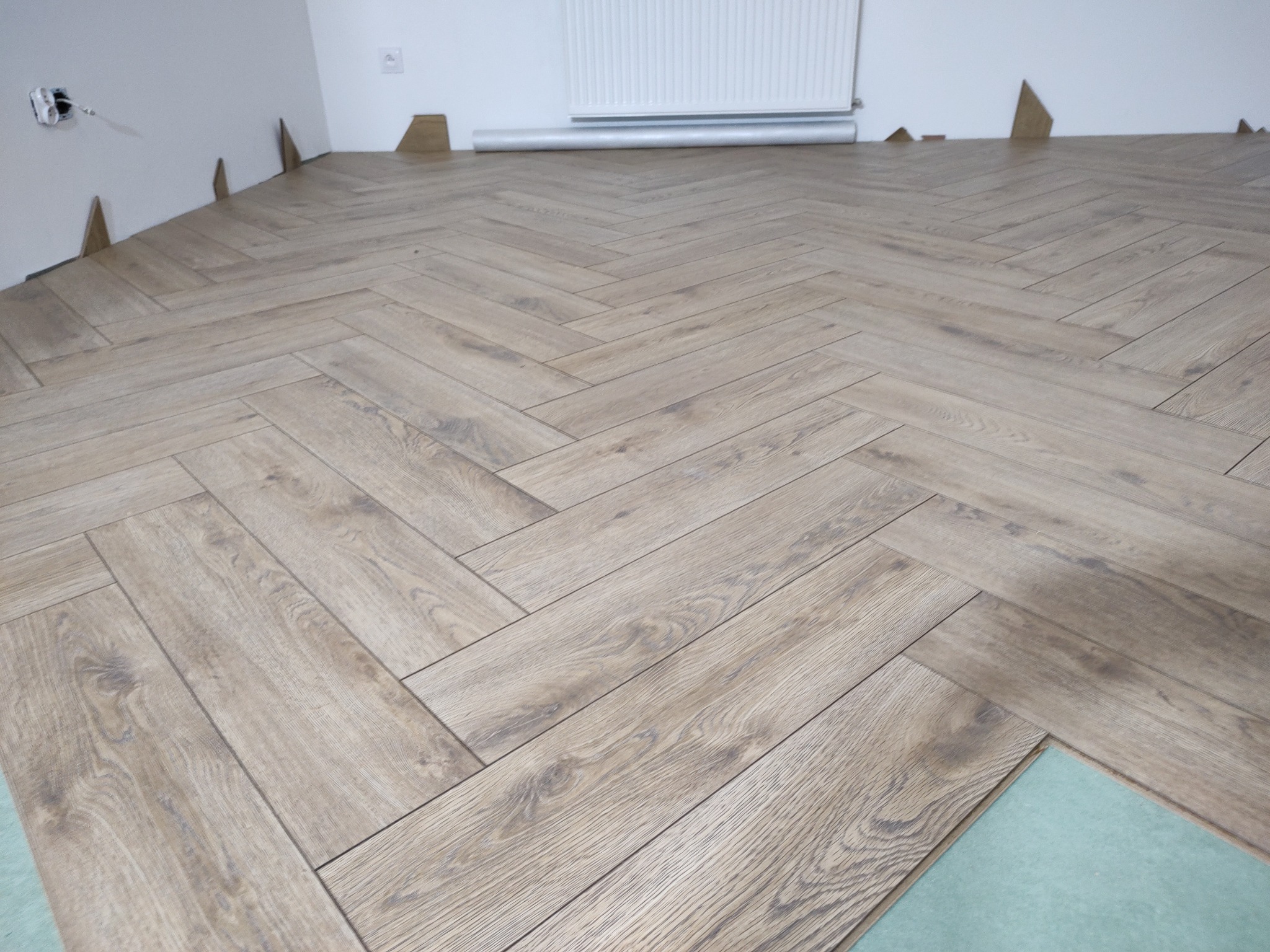 Ułożone panele w jodełkę, widoczny fragment podkładu. Częściowo wykończona podłoga w jasnych odcieniach brązu, listwy przypodłogowe nie zamontowane.